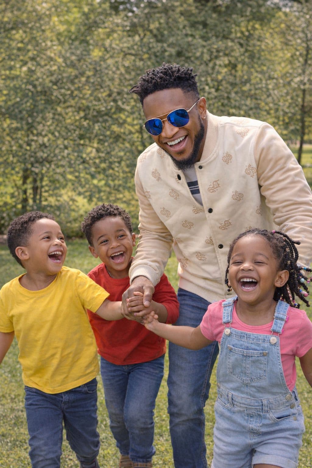 Smiling Black man and three laughing children holding hands while running in a sunny park.