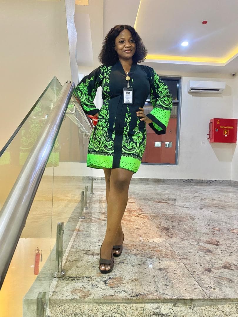 Woman in a black and green patterned dress posing by a glass staircase railing.