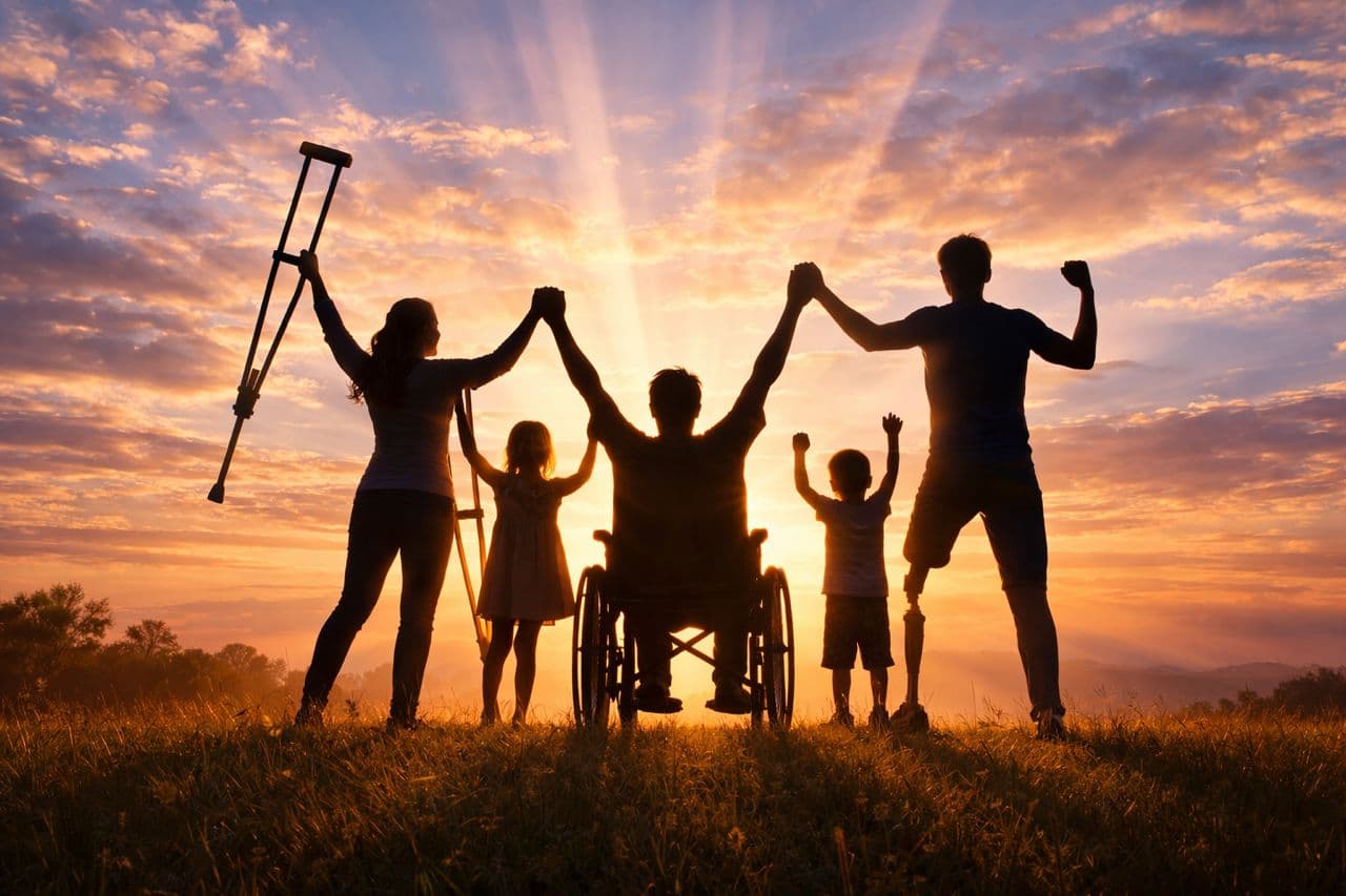 Silhouetted family with physical disabilities celebrating together against a radiant golden sunset in a field.