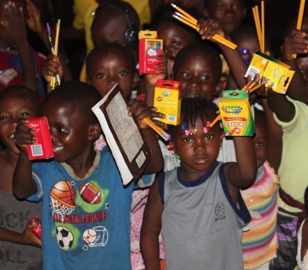 Smiling children hold up their new school supplies, including crayons, pencils, and a notebook.