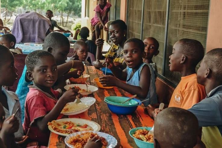 African children and a woman sharing a meal at a long wooden table outdoors.