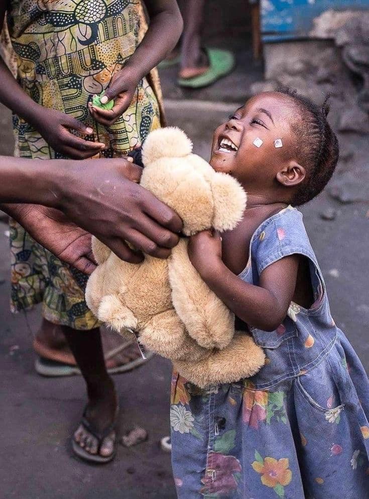 A joyful young girl laughs while receiving a tan teddy bear from outstretched hands.